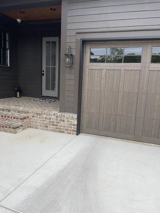 A single brown wood-look carriage door with a row of windows beside a brick stoop and dark siding