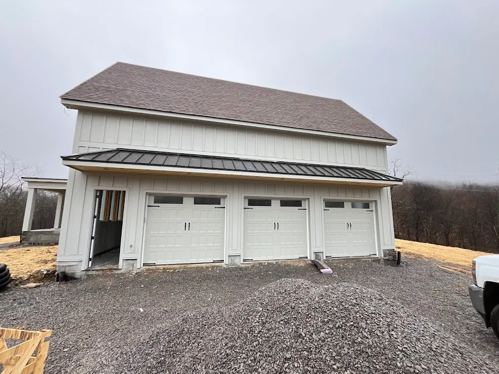 Three white carriage doors on a new board-and-batten farmhouse garage, gravel still in the drive